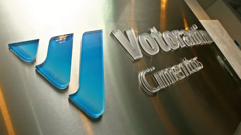 Logotipo da Votorantim Cimentos em parede metálica com acabamento polido e elementos em azul translúcido